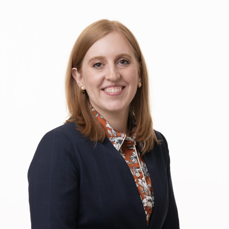 Smiling person with shoulder-length auburn hair wearing a navy blazer over a floral-patterned shirt, standing against a plain white background.