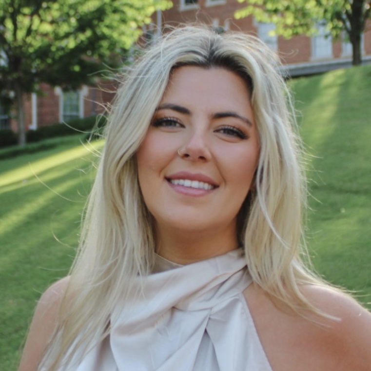 Smiling person with long blonde hair stands outdoors in sunlit park with green grass and trees. Wearing a light-colored top.