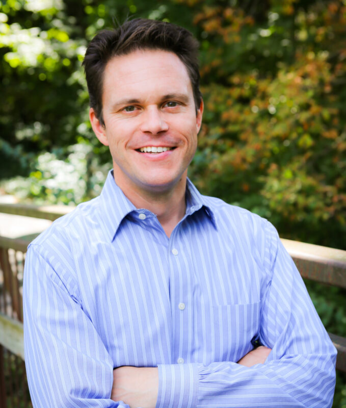 Man in a blue striped shirt smiling, standing outdoors with arms crossed, greenery in the background.