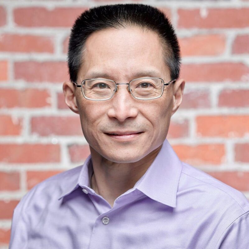 Man with glasses wearing a lavender shirt stands in front of a brick wall, looking at the camera.