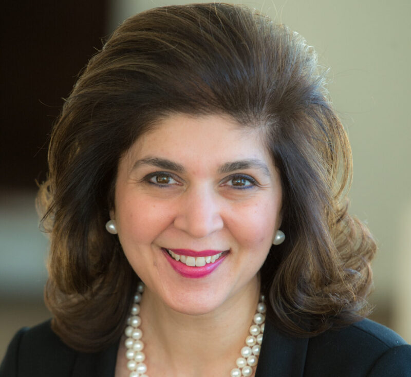 A woman with shoulder-length brown hair, wearing a black top and pearl necklace, smiles at the camera.