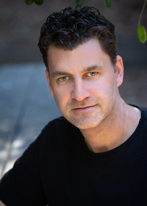 A man with curly dark hair and a black shirt looks forward with a soft expression, with blurred greenery in the background.