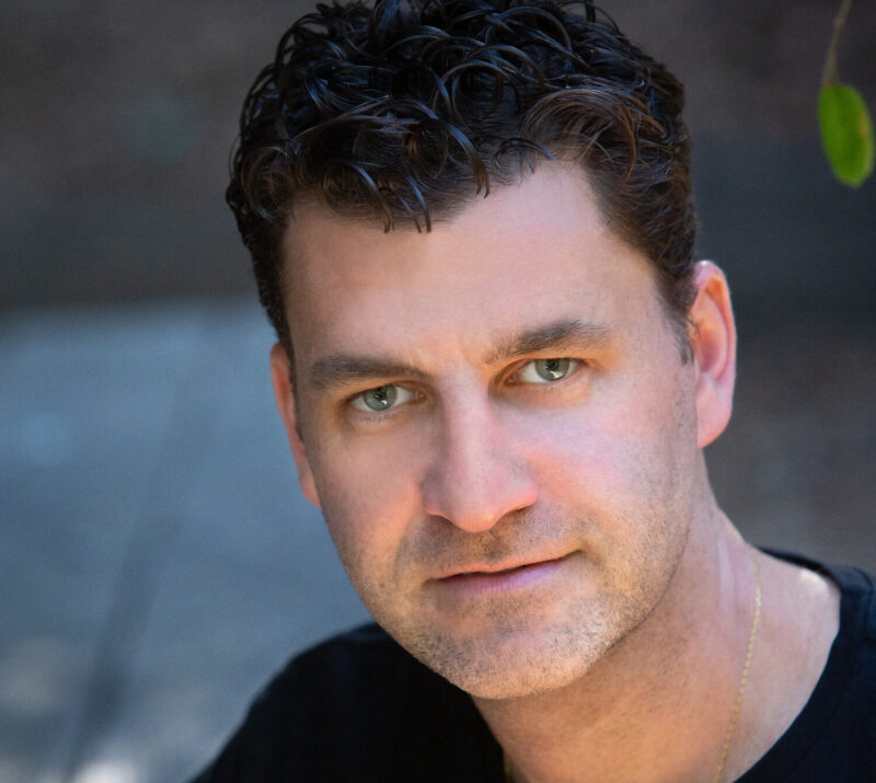 A man with curly dark hair and a black shirt looks forward with a soft expression, with blurred greenery in the background.