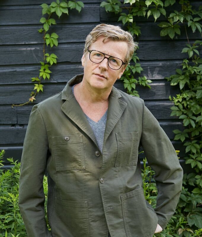 A person with glasses in a green jacket stands in front of a black wooden wall with vines.