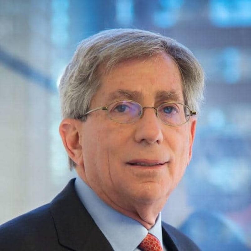 Man with glasses in a suit and tie, standing in front of a blurred background.