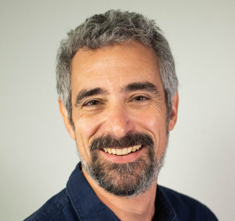 A man with gray hair and a beard smiles at the camera, wearing a dark blue shirt against a plain background.
