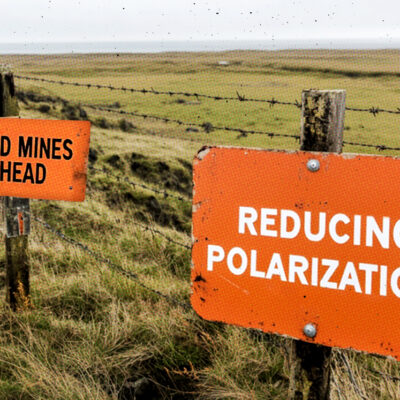 Two orange signs on a grassy landscape: one reads "LAND MINES AHEAD" and the other "REDUCING POLARIZATION." Both are attached to a barbed wire fence.