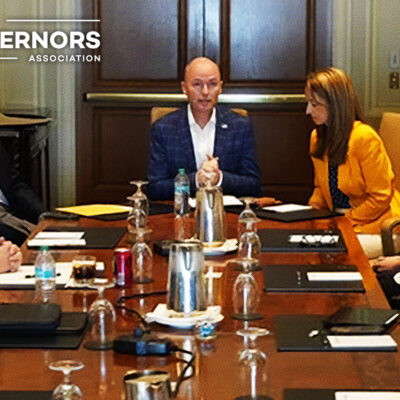 A group of people in business attire sit around a conference table discussing, with "National Governors Association" logo in the top left corner.