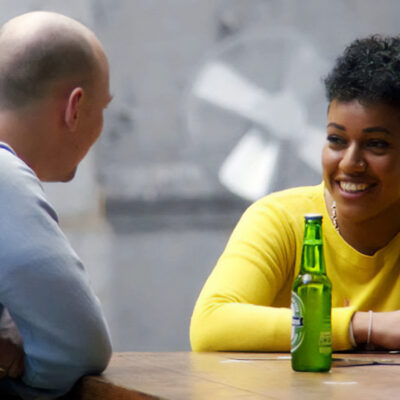 Two people sitting across from each other at a table with drinks. The woman in yellow is smiling, while the man in gray gestures with his hand.