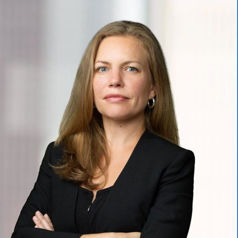 A woman with long blonde hair, wearing a black blazer, stands with her arms crossed and looks confidently at the camera.