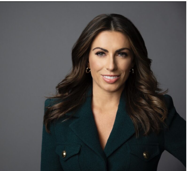 Smiling woman with long, wavy brown hair, wearing a dark green blazer, standing against a gray background.