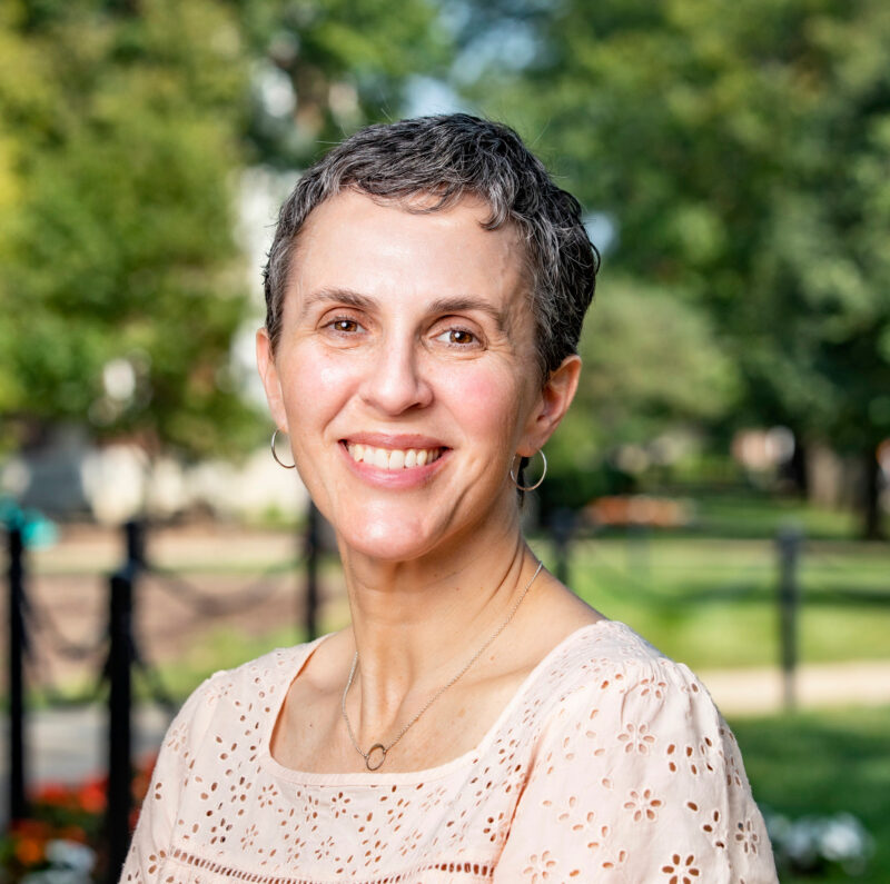 Smiling woman with short hair wearing a white blouse, standing outdoors with trees and greenery in the background.