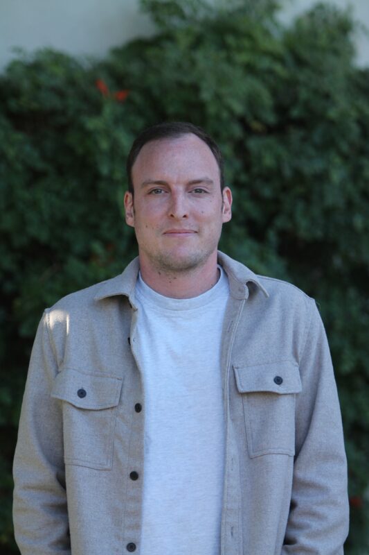 A person with short hair wearing a beige jacket and white shirt stands in front of green foliage, looking at the camera.