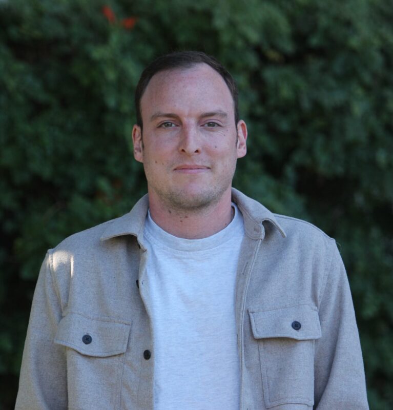 A person with short hair wearing a beige jacket and white shirt stands in front of green foliage, looking at the camera.