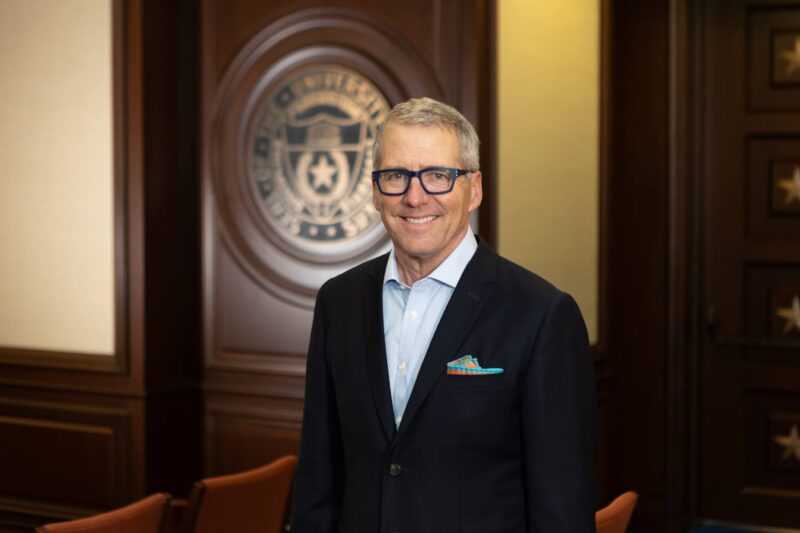 A man in a suit and glasses stands smiling in a wood-paneled room with a large emblem on the wall behind him.