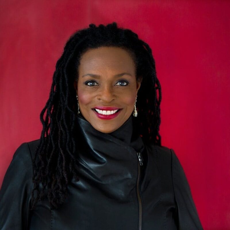 A woman with long braided hair wearing a black jacket smiles against a red background.