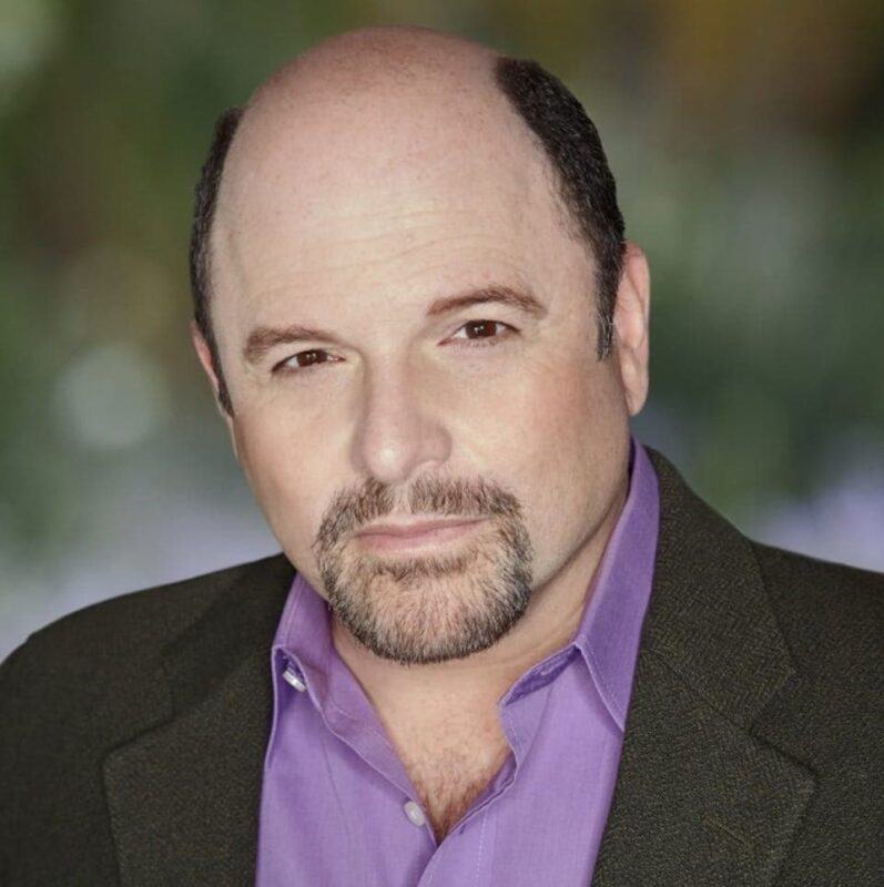 Man with a goatee and bald head, wearing a purple shirt and dark blazer, looking into the camera against a blurred background.