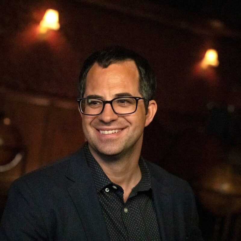 Smiling man in glasses and a suit jacket with a polka dot shirt, standing indoors with dim lighting and wall sconces in the background.