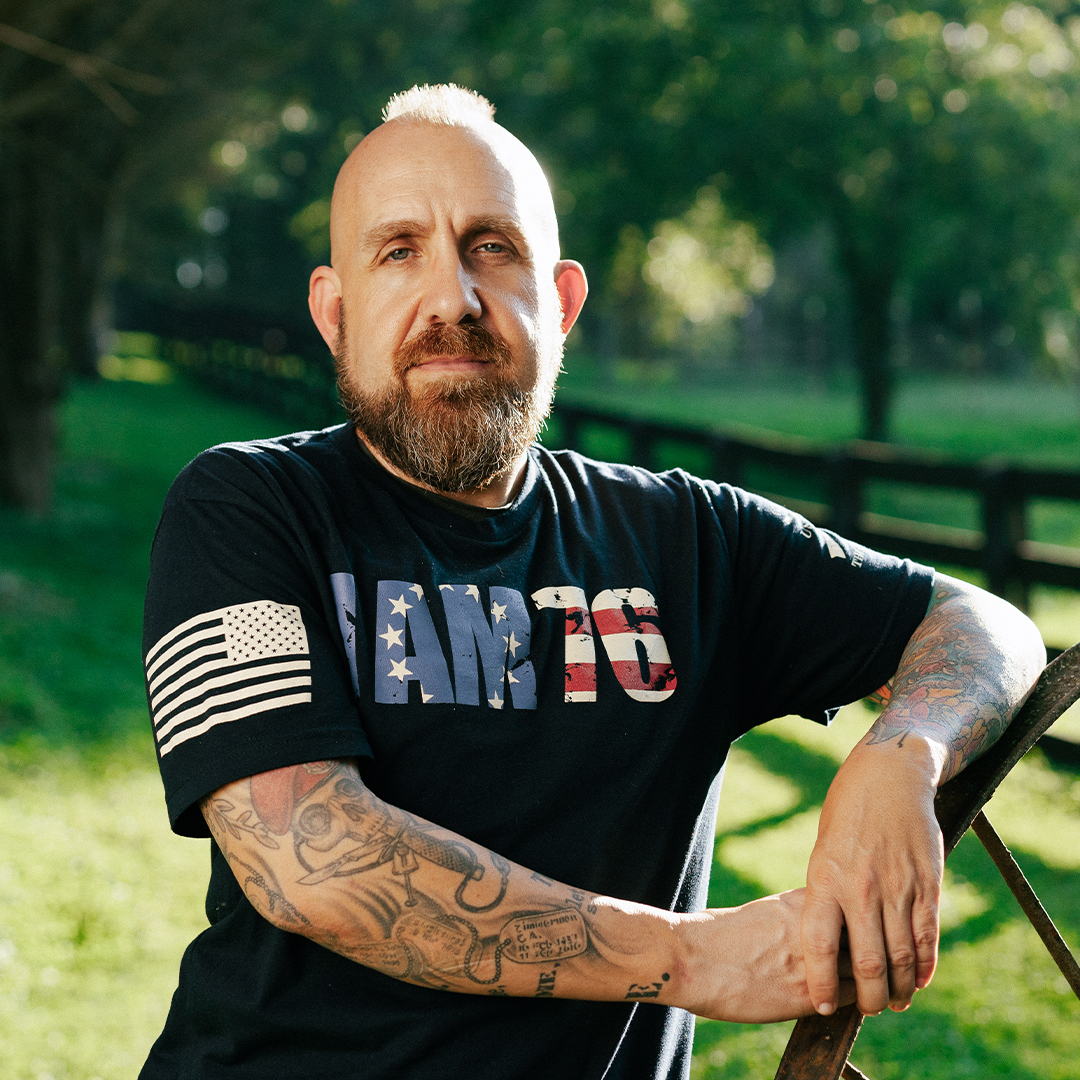 A bearded man with tattoos and a mohawk stands outdoors, wearing a black T-shirt with an American flag design. Trees and a fence are in the background.