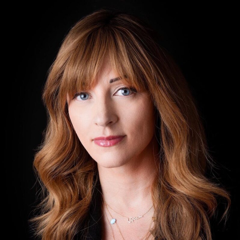 Woman with long, wavy brown hair and bangs, wearing a necklace, against a black background.