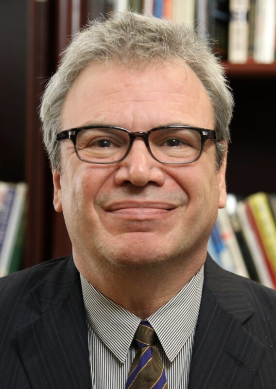 Man with glasses, gray hair, and a suit, smiling slightly in front of a bookshelf.