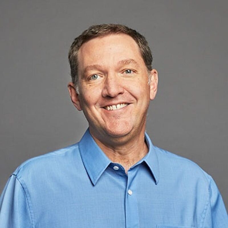 Man in a blue shirt smiling against a gray background.