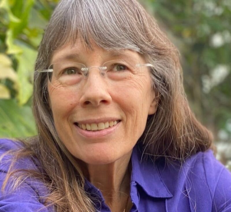 Smiling woman with long gray hair and glasses in a purple shirt, outdoors with greenery in the background.