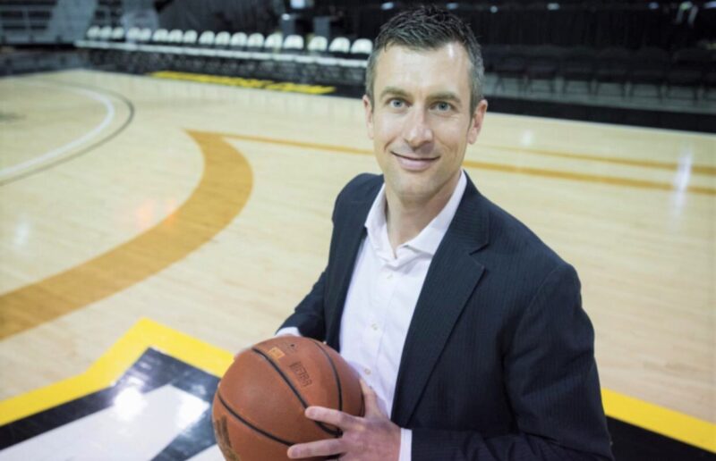 A man in a suit holding a basketball stands on a basketball court, facing the camera with a smile.