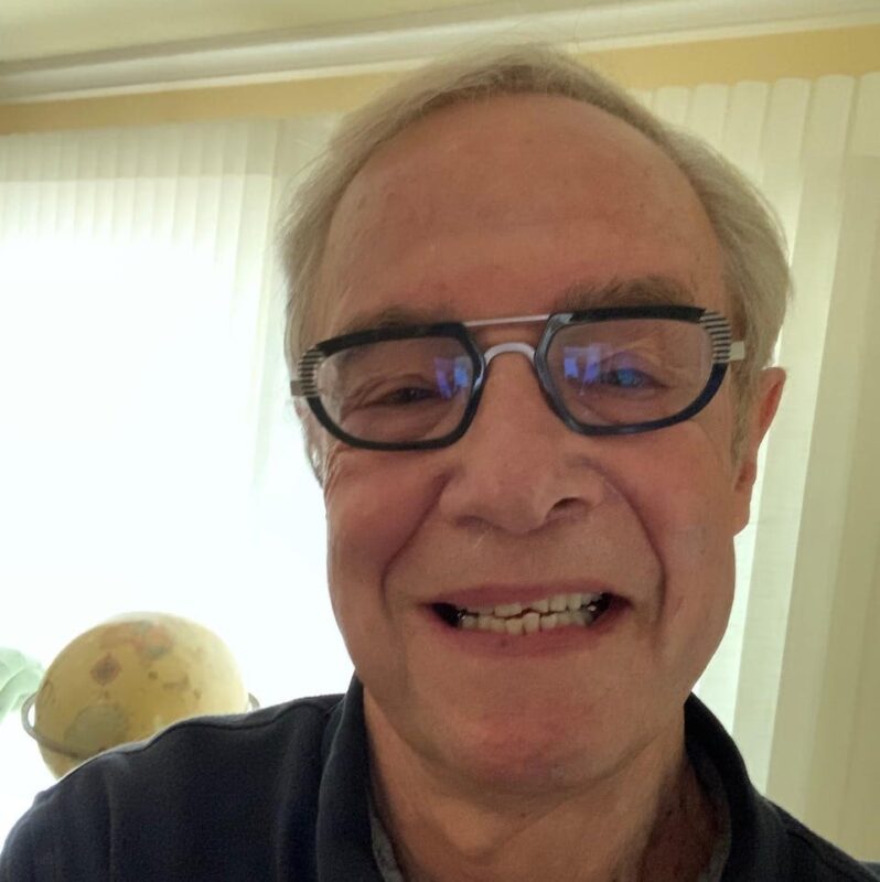 Elderly man wearing glasses smiling in a warmly lit room with sheer curtains and a globe in the background.