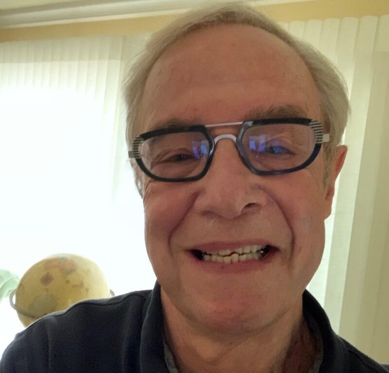 Elderly man wearing glasses smiling in a warmly lit room with sheer curtains and a globe in the background.