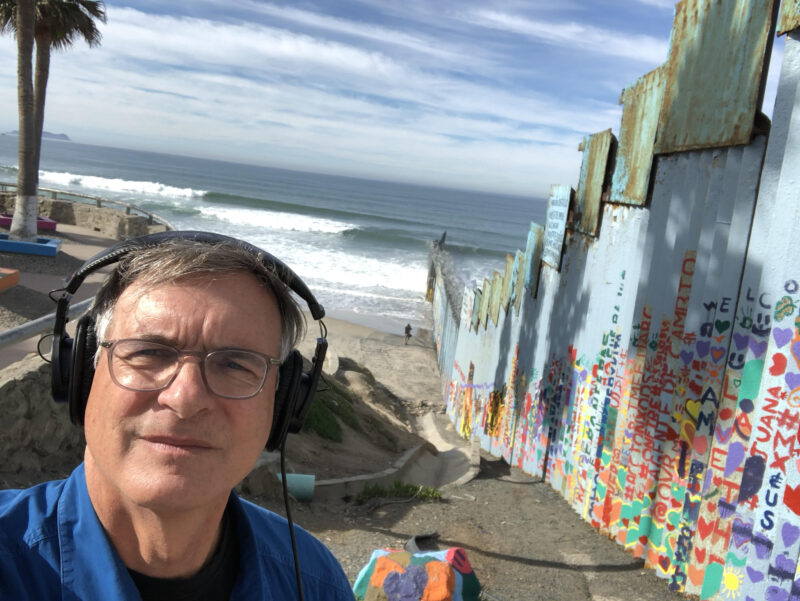 A person wearing headphones stands near a colorful, graffiti-covered wall by the ocean, with waves and a cloudy sky in the background.