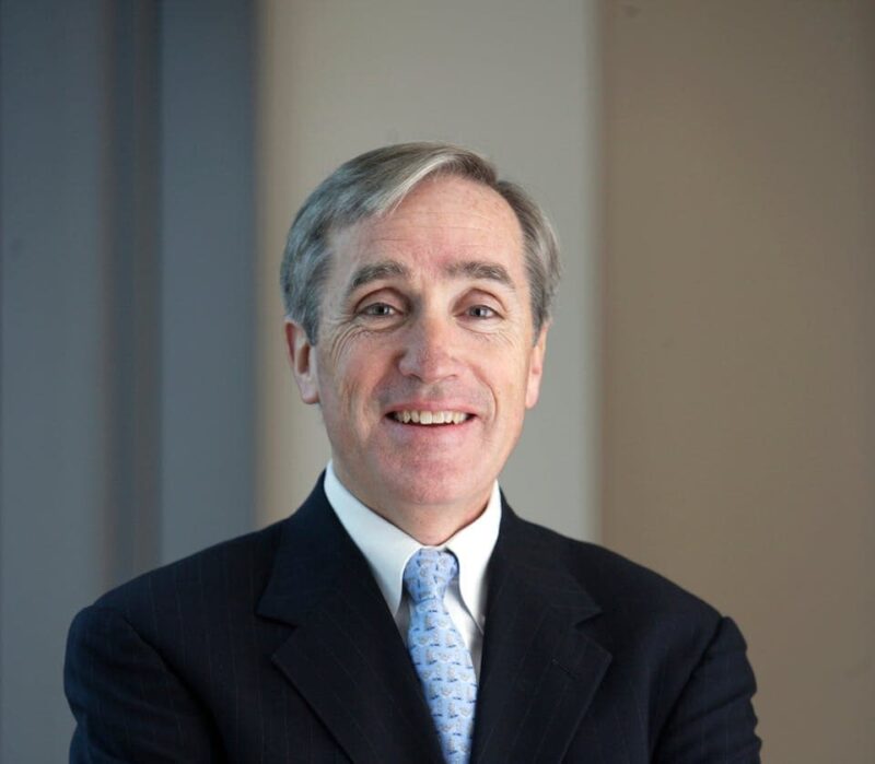 A smiling person with gray hair wearing a dark suit and blue tie stands in front of a beige and blue background.