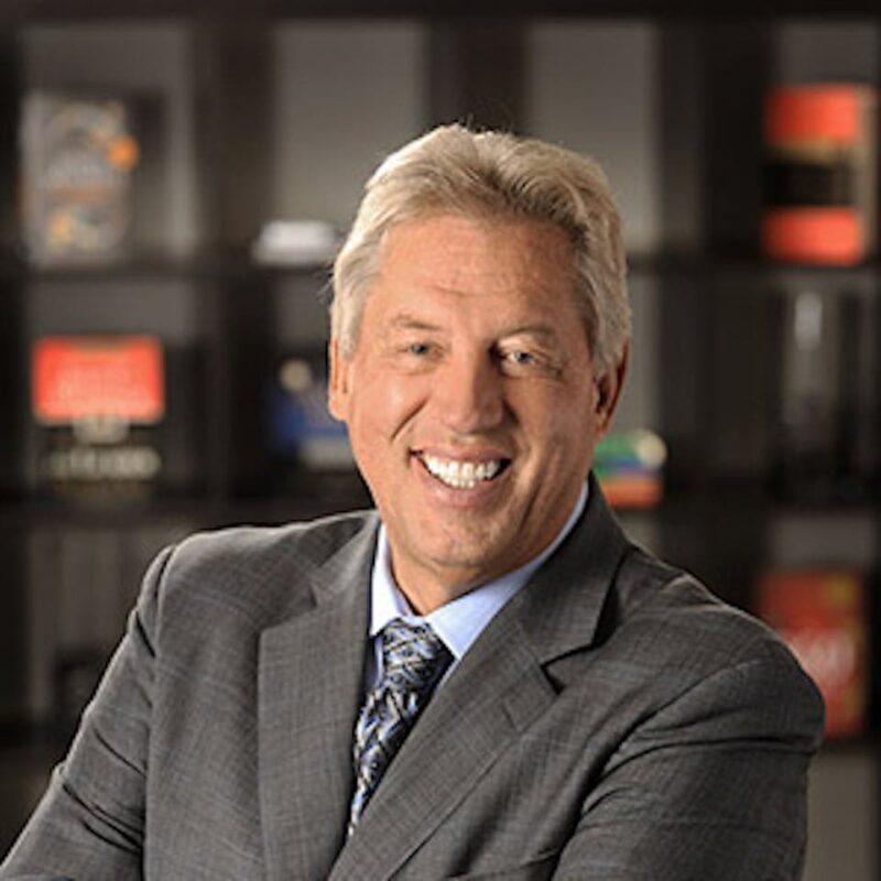 Smiling person with gray hair in a suit and tie, standing in front of a bookshelf.