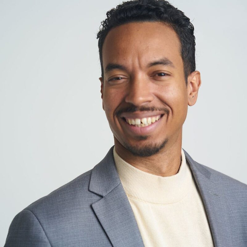 Smiling man in a gray suit jacket and cream turtleneck, with short curly hair, against a plain light background.