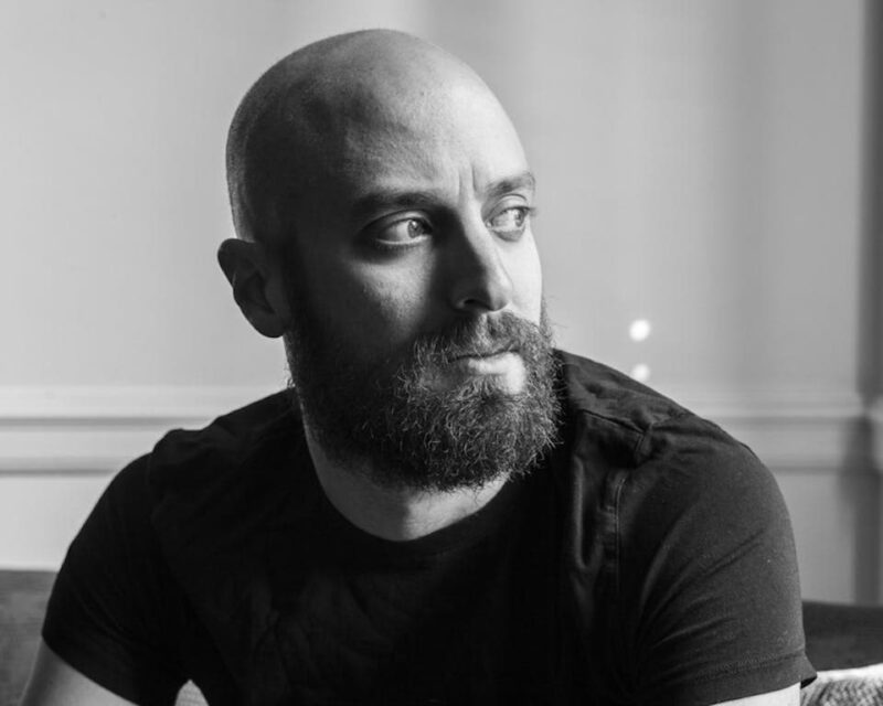 Black and white image of a bearded man with a shaved head, wearing a dark T-shirt, looking to his right.