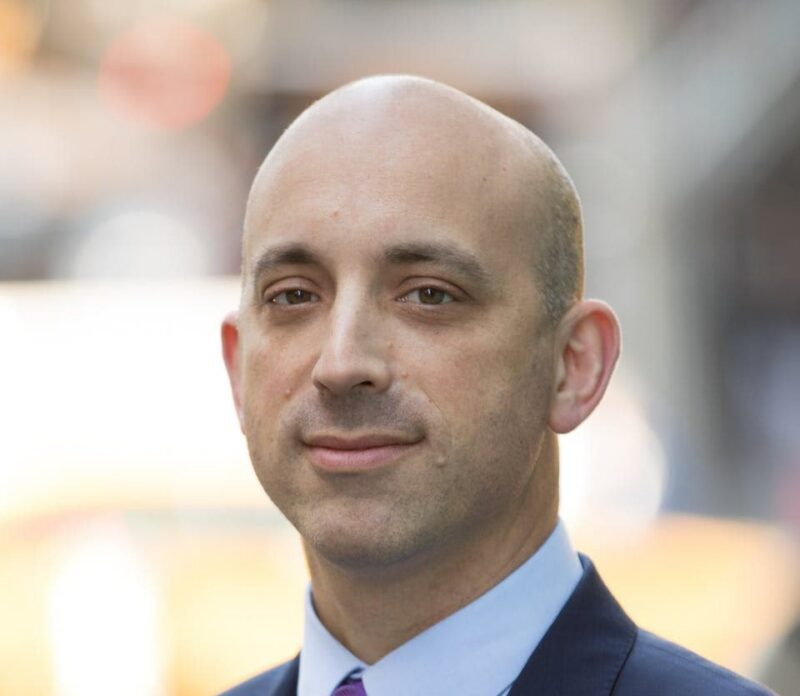 A bald man in a suit standing outdoors with blurred city lights in the background.