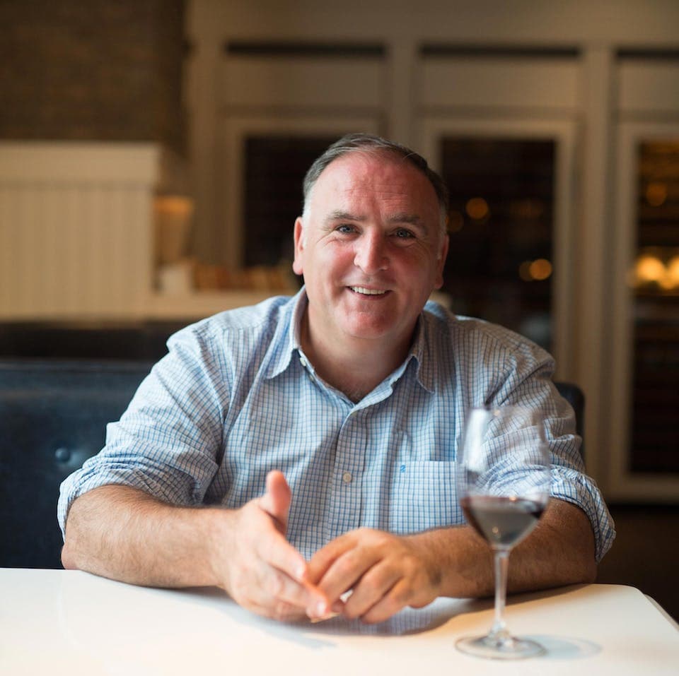 Man in a checkered shirt sitting at a table with a glass of red wine, smiling at the camera, in a warmly lit restaurant.