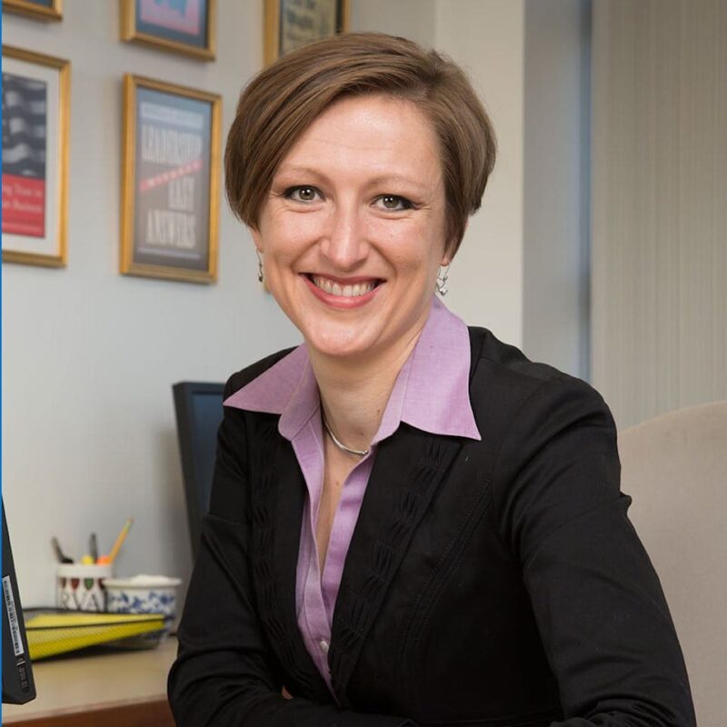 Smiling person with short hair in a black blazer and purple shirt, sitting at a desk with framed certificates in the background.