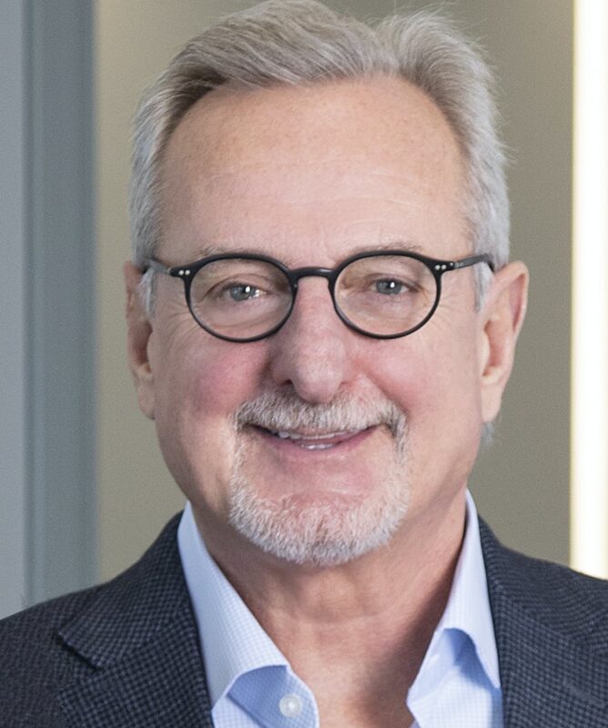 Man with glasses, a gray beard, and short gray hair, wearing a blue shirt and a dark blazer, smiles in a professional setting.