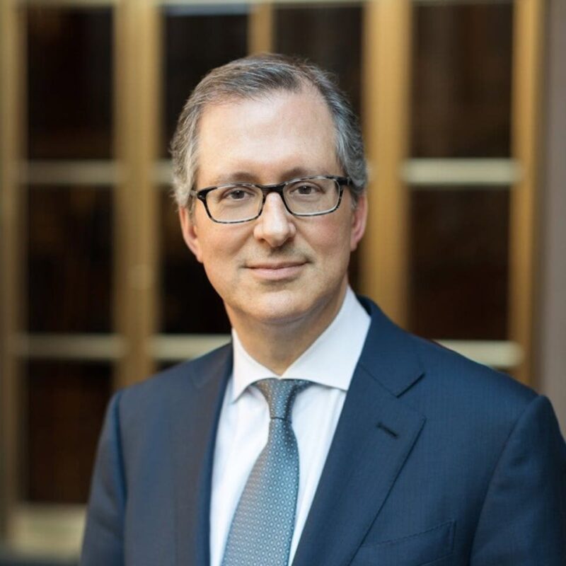 A person wearing glasses and a business suit stands in front of glass-paneled doors, looking at the camera.