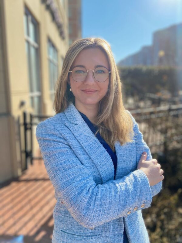 A woman in a light blue blazer and glasses stands outdoors, smiling with her arms crossed. She is in front of a building on a sunny day.