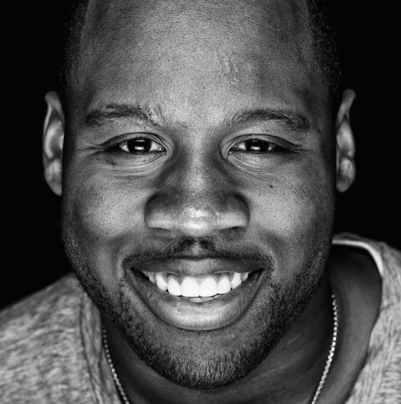 Smiling man in grayscale photo, wearing a light-colored shirt and a simple chain necklace against a dark background.