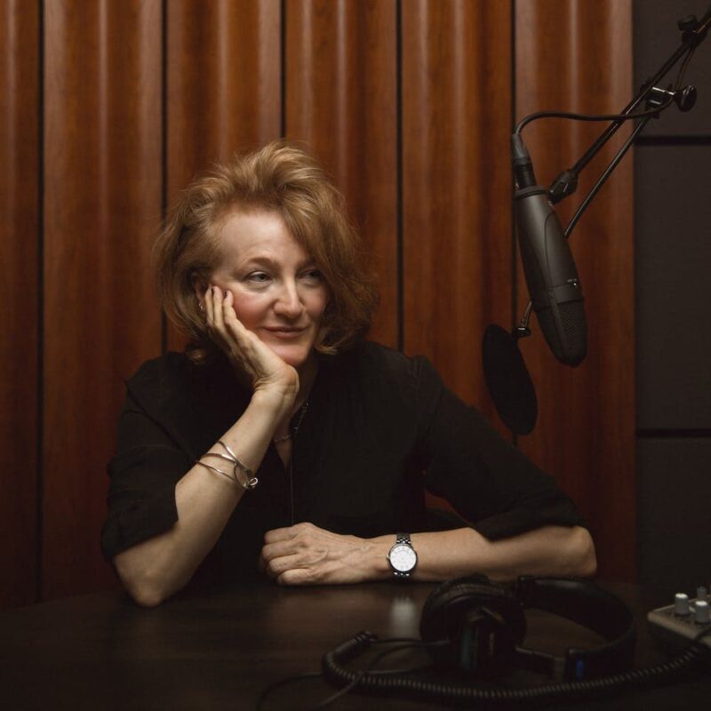 Person sitting at a table with a microphone, wearing a black shirt, watch, and bracelets, leaning on one hand, in front of a wooden background.