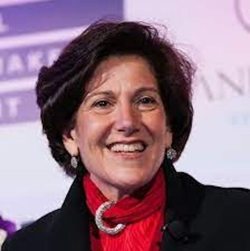 A person with short dark hair smiles, wearing a black jacket and red scarf. The background is partially blurred with purple and white hues.