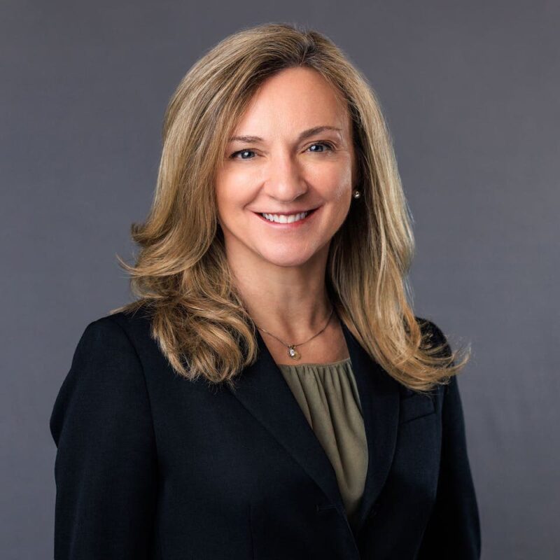 Smiling woman with long blonde hair in a black blazer and olive blouse, standing against a gray background.