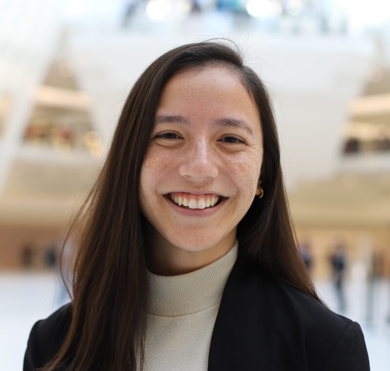 A person with long hair smiling, wearing a black blazer and light-colored top, in a bright indoor setting.