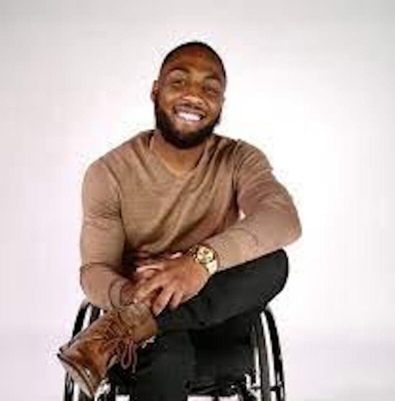 Smiling man in a wheelchair, wearing a brown long-sleeve shirt and brown shoes, sitting against a plain white background.