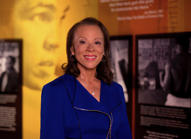 Smiling woman in a blue outfit stands in front of a wall with images and text.