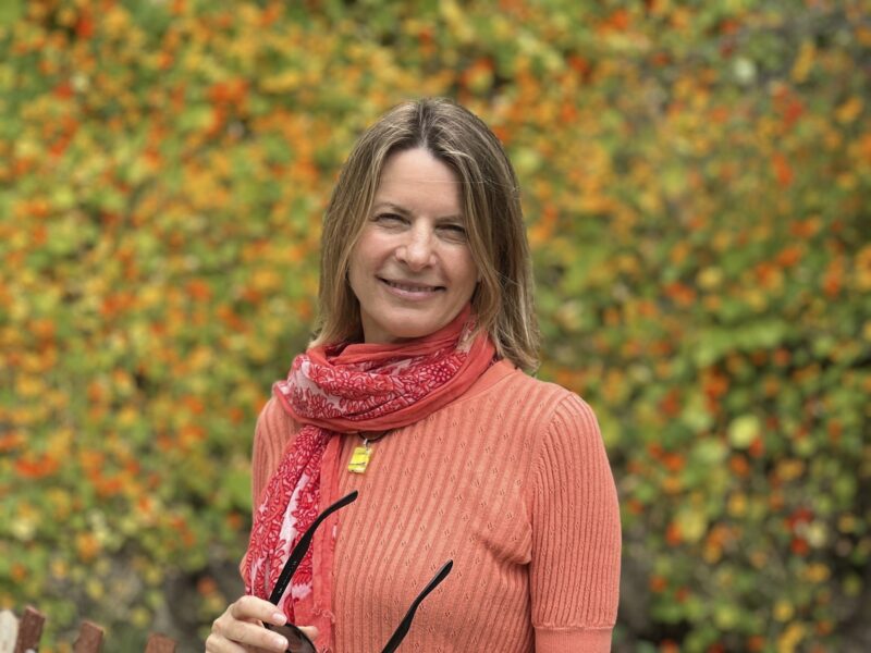 Woman smiling outdoors in front of a vibrant autumn background, wearing an orange sweater and red scarf, holding glasses.