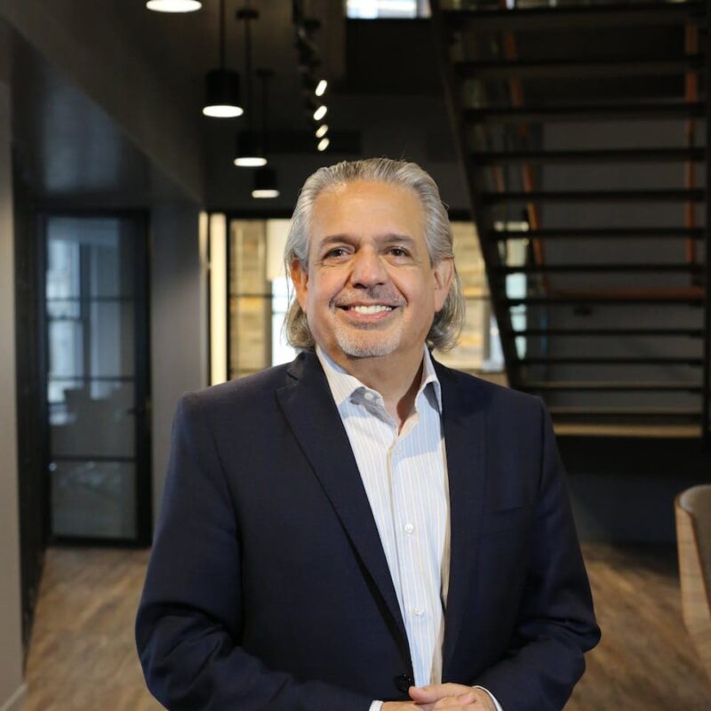 Smiling man with gray hair in a dark suit stands in a modern office hallway with stairs and hanging lights in the background.